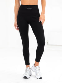 A model wearing high-waisted black active leggings with a small blakely brand logo on the waistband, paired with white sneakers.